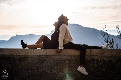 In Grenoble, France, a couple sits back to back on a wall at a lookout point, holding hands and touching heads for a portrait.