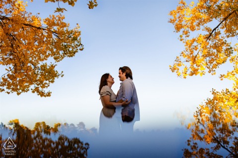 At Washington Park in Denver, Colorado, an engaged couple stands face to face in profile, framed by autumn leaves and reflections, with a blue sky in the background for their portrait.