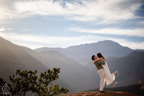 At Garden of the Gods in Colorado Springs, Colorado, the groom-to-be lifts his fiancée as they pose in a dancing embrace with mountain views in the background for their engagement portrait.