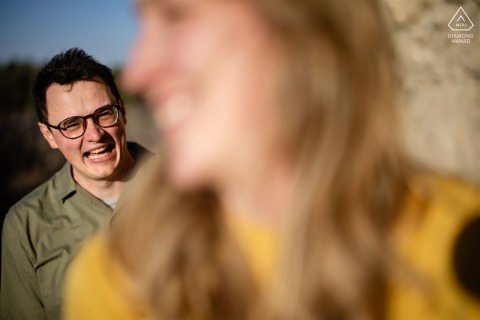 At Monument Rock in Monument, Colorado, the groom-to-be laughs in focus while his fiancée appears out of focus in the foreground during their portrait session.