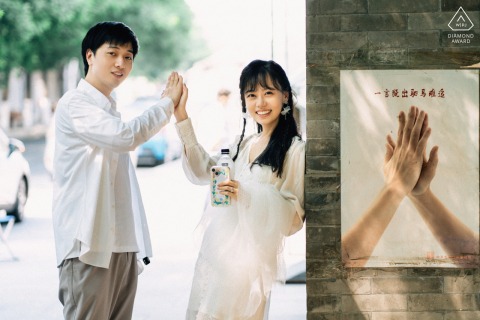Sanlihe Engagement Portrait: Couple High-Fiving Before Wall, Mirroring Nearby Photo At Sanlihe in Beijing, China, a couple poses for a portrait as they high-five in front of a wall, mirroring a photograph displayed nearby.