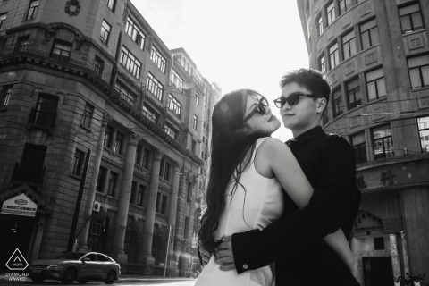 At the Bund in Shanghai, China, a couple wearing sunglasses and formal attire embraces in bright sunlight, posing for a portrait with vintage Hollywood style.