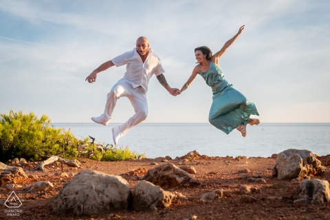 In Bandol, Var, Provence, France, a couple holds hands and jumps by the sea, clicking their heels together in playful, energetic poses for a portrait.