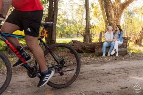 In Parque Villarino, Argentina, a couple sits together on a park bench during a pre-wedding session as a bicyclist passes by in the foreground.