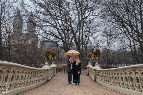 Central Park Pre-Wedding Portrait: Couple Under Umbrella on Footbridge In Central Park, New York, a couple stands together under an umbrella at the center of a footbridge, posed for a portrait a few days before their wedding.
