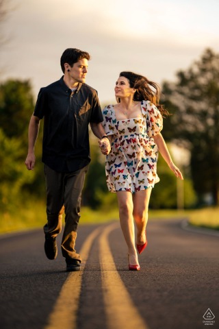 In Doylestown, Philadelphia, an engaged couple poses running hand in hand down a road marked with yellow stripes, framed by the long stretch of pavement ahead in a cinematic-style portrait.