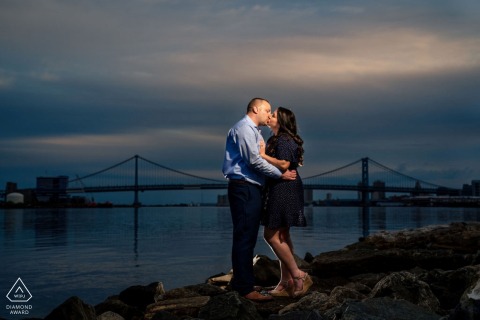 At the Philadelphia Navy Yard in Pennsylvania, an engaged couple shares a kiss at twilight, posed among rocks with a quiet bridge over the water in the background, illuminated by the photographer’s expert use of on-location lighting.