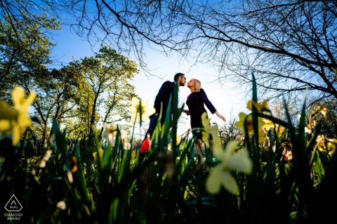 In Philadelphia, a couple leans in through tall grass and flowers, about to kiss, with trees and a blue sky above, captured in a portrait that highlights the photographer’s mastery of location lighting.