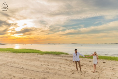 At Millway Beach in Barnstable, Massachusetts, a couple walks hand in hand along the shoreline during sunset, against the horizon in a wide, scene-setting shot.