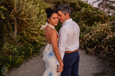 On a narrow path at The Knob in Falmouth, Massachusetts, an engaged couple pauses with eyes closed and foreheads touching, bathed in warm coastal light during a portrait session.
