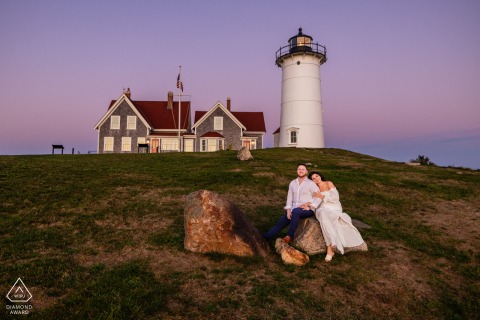 Below Nobska Lighthouse in Falmouth, Massachusetts, a couple sits together between boulders in soft twilight as evening light settles over the scene.