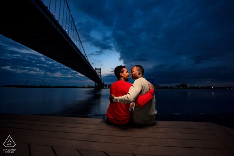 At Race Street Pier in Philadelphia, a couple holds each other close during twilight, framed by a bridge extending over shimmering water in a portrait that highlights the photographer’s mastery of on-location lighting.