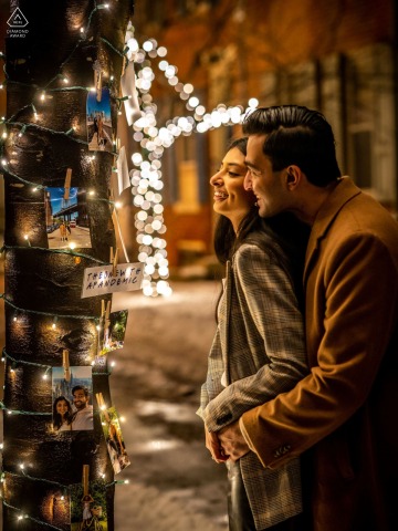 Philadelphia Night Engagement Portrait: Embrace on Addison Street with City Lights On Addison Street in Philadelphia, a man wraps his arms around his fiancée from behind for a night portrait, with illuminated trees and city lights in the background.