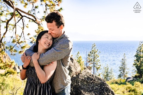 South Lake Tahoe Engagement Portrait: Embrace by the Lake At South Lake Tahoe, California, a man wraps his arms around his fiancée from behind as she smiles, with the lake visible in the background during their engagement portrait session.