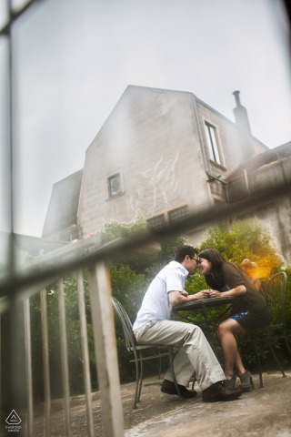 Seated at a patio table in San Francisco, California, two people gaze at each other affectionately in a vertical engagement portrait captured from a low, tilted angle.