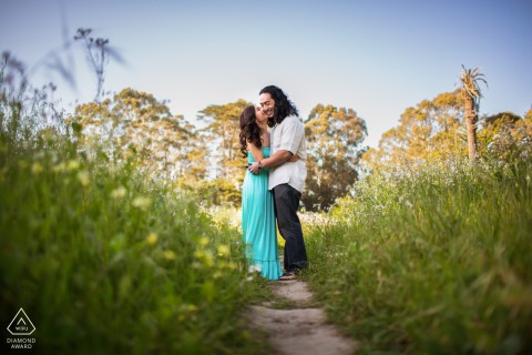 An engaged couple stands face to face in profile on a nature path bordered by green grass, captured from a low angle during a portrait session in Santa Cruz, California.