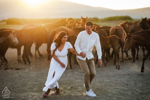 Engaged Pair Dancing at Sunset in Kayseri Horse Corral An engaged couple dances in a horse corral during a photo session in Kayseri, Turkey, as the sun sets behind them and several horses kick up dust in the background.