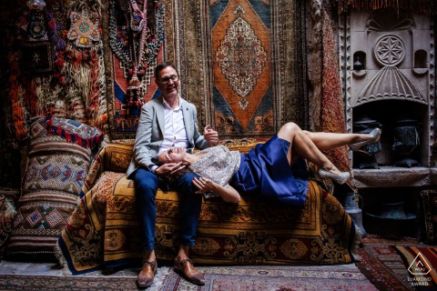 An engaged couple poses on a couch inside a carpet vendor’s shop during a pre-wedding photo session in Cappadocia, Göreme, Nevşehir, Turkey, with natural light streaming from above.