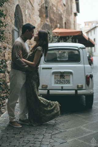 An engaged couple kisses passionately on a cobblestone alleyway in Italy, with a vintage small car parked behind them.