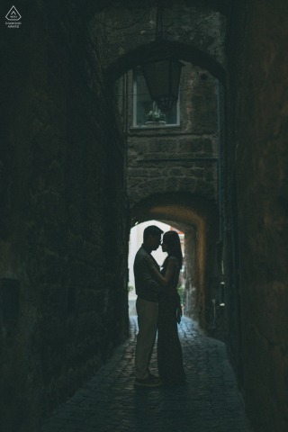 A silhouetted engaged couple faces each other in a tall, dramatic vertical portrait in a small town in Italy.