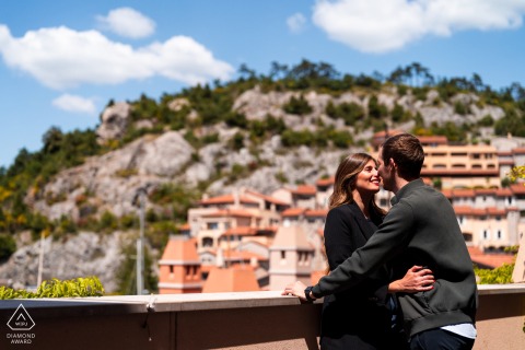 An engaged couple hugs in sunlight with a hillside of dwellings visible behind them in Portopiccolo, Sistiana, Trieste, Italy, during a portrait session.
