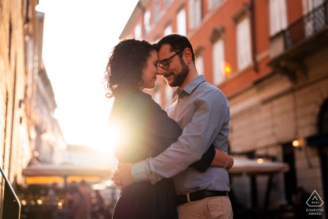 An engaged couple hugs in the warm glow of sunset as sunlight bursts between buildings behind them in Trieste, Italy, during a portrait session.
