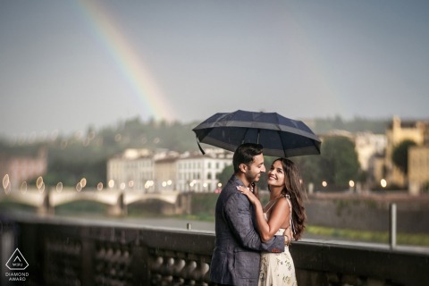 A couple shares a joyful embrace under an umbrella on a Florence street, framed off-center, with a vibrant rainbow arching behind them, adding a colorful touch to the scene.