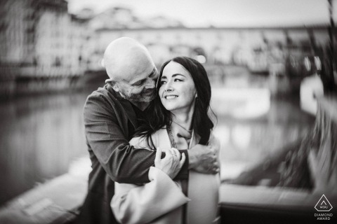 A couple shares an intimate embrace by the river in Florence, captured in black and white with a Petzval lens, creating characteristic swirly bokeh for an artistic, romantic effect.
