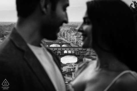 In this black and white photo, a softly focused couple in the foreground frames the iconic Ponte Vecchio, adding a romantic, dreamy atmosphere to a scenic Florence cityscape.