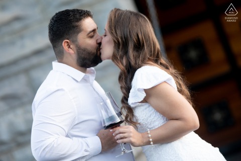 Couple standing and sharing a kiss beside stones of an old world building at Cave Brandalize in Monte Belo do Sul, holding glasses of rich red wine.