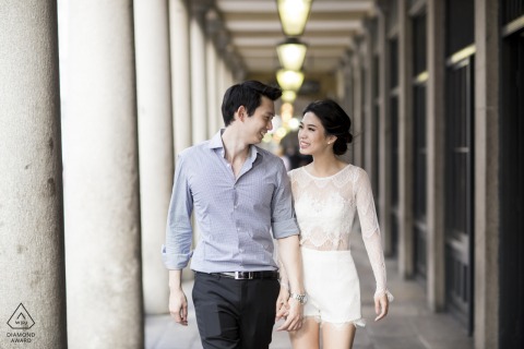 Couple walking side by side in London, exchanging warm smiles as they look at each other, moving past classic porch columns in a charming, urban engagement portrait.