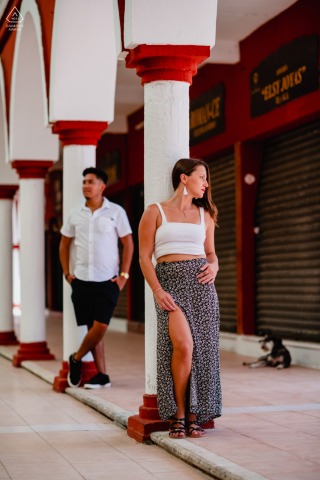 Couple stands apart, each leaning against separate columns on a porch at a Valladolid marketplace in the early morning, quietly sharing a thoughtful connection in the soft light.