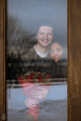 Newly engaged couple stands together at Kondiaronk Belvedere in Montreal, smiling through a glass doorway and holding a bouquet of roses.