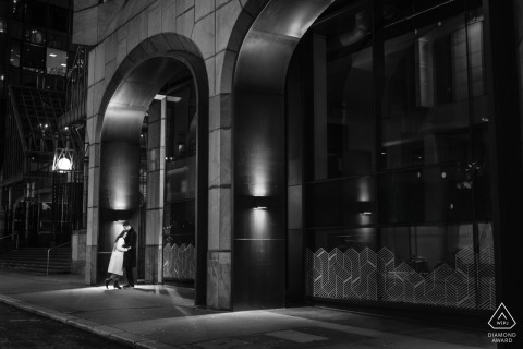 Couple stands together on a city sidewalk at night in the City of London, captured in a black and white portrait that highlights the urban lights and nighttime atmosphere surrounding them.