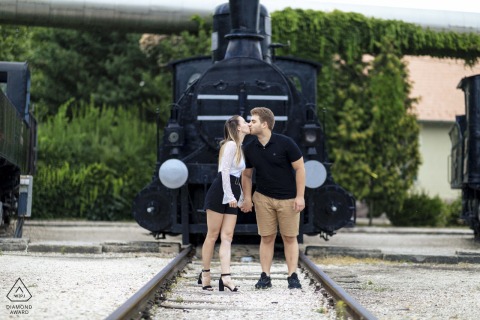 Couple shares a kiss while holding hands on railway tracks in Budapest, with an old locomotive standing quietly behind them, adding a nostalgic and romantic atmosphere to the scene.
