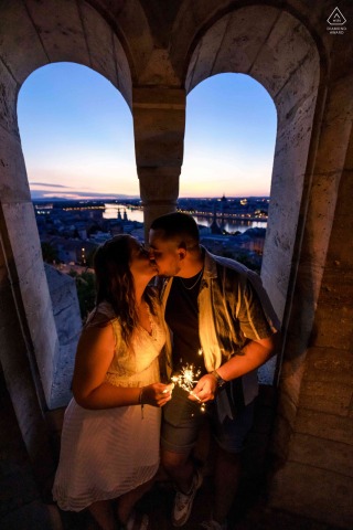 Couple shares a kiss at dusk beneath ancient stone arches inside an old world castle tower in Budapest, their sparklers glowing warmly against the city lights twinkling below.