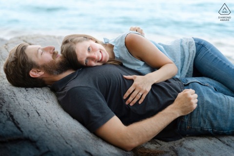 In Freeport, Maine, a couple lies on coastal rocks with her head resting on his chest. She smiles at the camera, displaying her engagement ring, creating a warm, intimate scene.