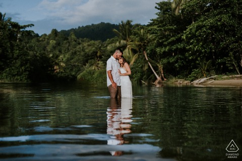 Engagement portrait at Las Cuevas Beach, Trinidad, features the couple standing in the middle of a tranquil river flowing toward the sea behind them, illuminated by golden hour sunlight.