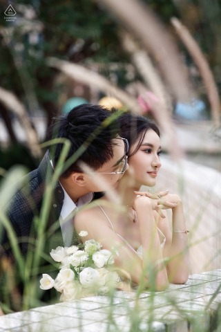 Amidst the greenery on the Bund in Shanghai, he gently kisses her, their intimate connection softly revealed through wispy grass blades in an artful, nature-inspired engagement portrait.