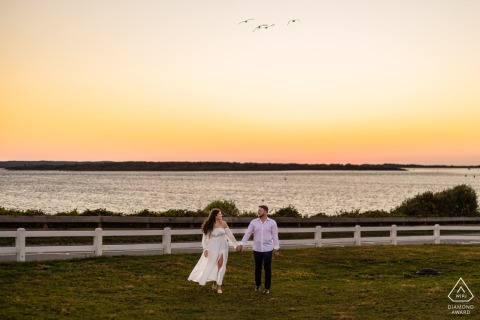 A couple walks hand in hand along the shoreline near Nobska Lighthouse in Falmouth, MA at sunset, surrounded by soft pastel skies and calm light, capturing timeless Cape Cod romance.
