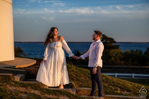 Bathed in soft evening light near Nobska Lighthouse, Falmouth, MA, the couple holds hands with the ocean behind them, reflecting the calm and intimacy of a Cape Cod engagement session.