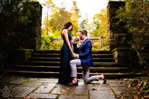 He kneels on the stone path at Hunting Hill Mansion, holding her hand as autumn light surrounds them. Center framed and backlit, the couple is framed by stone columns behind them.