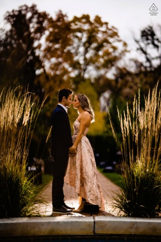 The couple stands hand in hand on a garden path at Longwood Gardens in Philadelphia, backlit by the sun and framed by tall grasses and warm evening light in a tall vertical portrait.