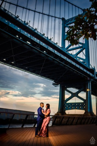 The couple stands close beneath the bridge at Race Street Pier in Philadelphia, embracing as the city and river settle into the evening light, framed in a tall, dramatic vertical shot with a striking sky.