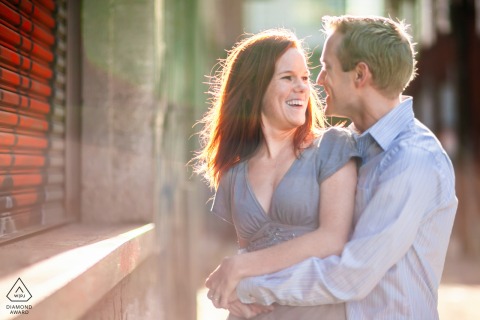 On a sunlit urban sidewalk in San Francisco, the couple stands outside a shop, backlit and smiling at each other, capturing a lively and affectionate engagement portrait in the city.