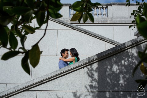 At UC Berkeley, California, a couple embraces on intersecting stone staircases outdoors. Sunlight bathes the historic campus architecture, lending the engagement portrait an inviting, romantic, old world charm.