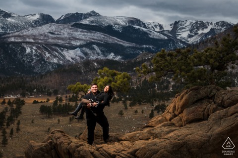 Future Groom Playfully Hoists Bride Into His Arms Amidst Mountains At Rocky Mountain National Park The future groom playfully hoists his bride into his arms amidst the mountains of Colorado at Rocky Mountain National Park, Estes Park, creating a spirited and energetic engagement image.