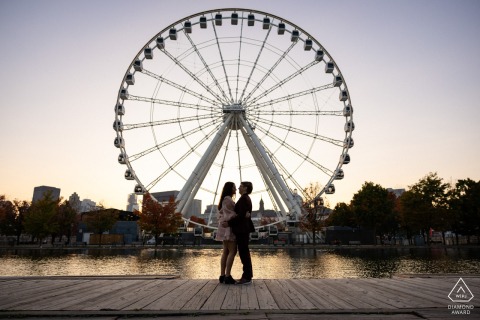 he striking silhouette of an engaged couple is captured in a romantic embrace in front of the grand ferris wheel at La Grande Roue in Montreal, Quebec, during sunset.
