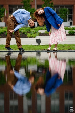 An engaged couple leans over playfully to view their upside-down reflections in a still water pool, creating a unique and artistic engagement portrait in the city of Montreal, Quebec.