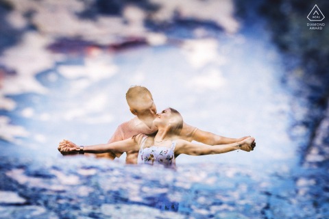 A couple shares a tender interaction at Borgo di Corsignano, Tuscany, reflected beautifully in a rain puddle after a summer shower, with their joyful pose appearing like a graceful dance.